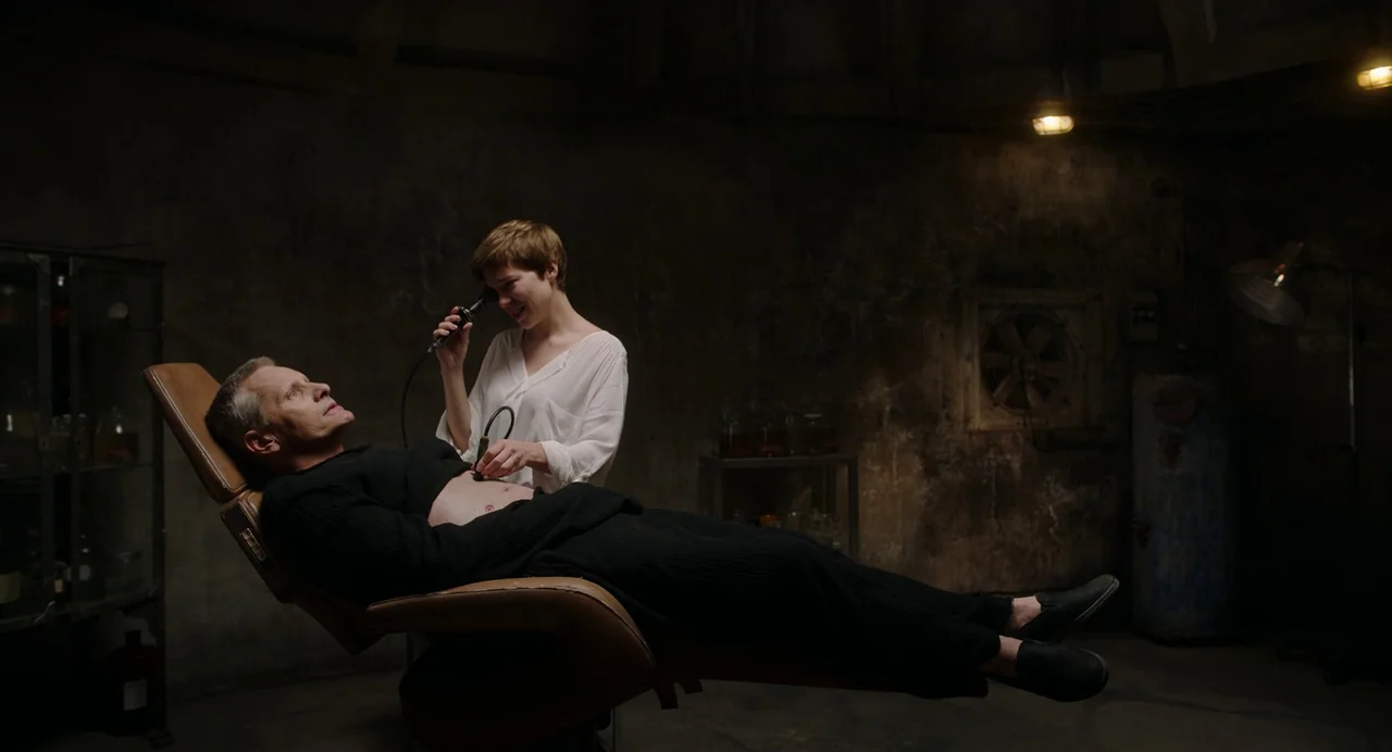 Crimes of the Future (2022) movie still: medium — An industrial-looking medical room features dark textured walls and a ventilation fan. A woman in…