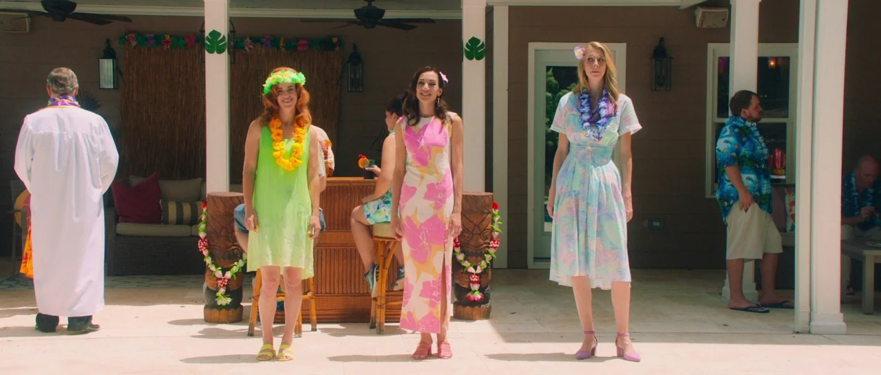Greener Grass (2019) movie still: fullbody — A sunlit patio features a tiki-themed bar and white columns supporting a roof. Three women stand side…