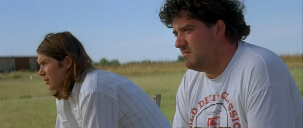 Shotgun Stories (2007) movie still: medium — Two men sit in a large, open grassy field under a bright clear sky. The pair look forward with neutral…