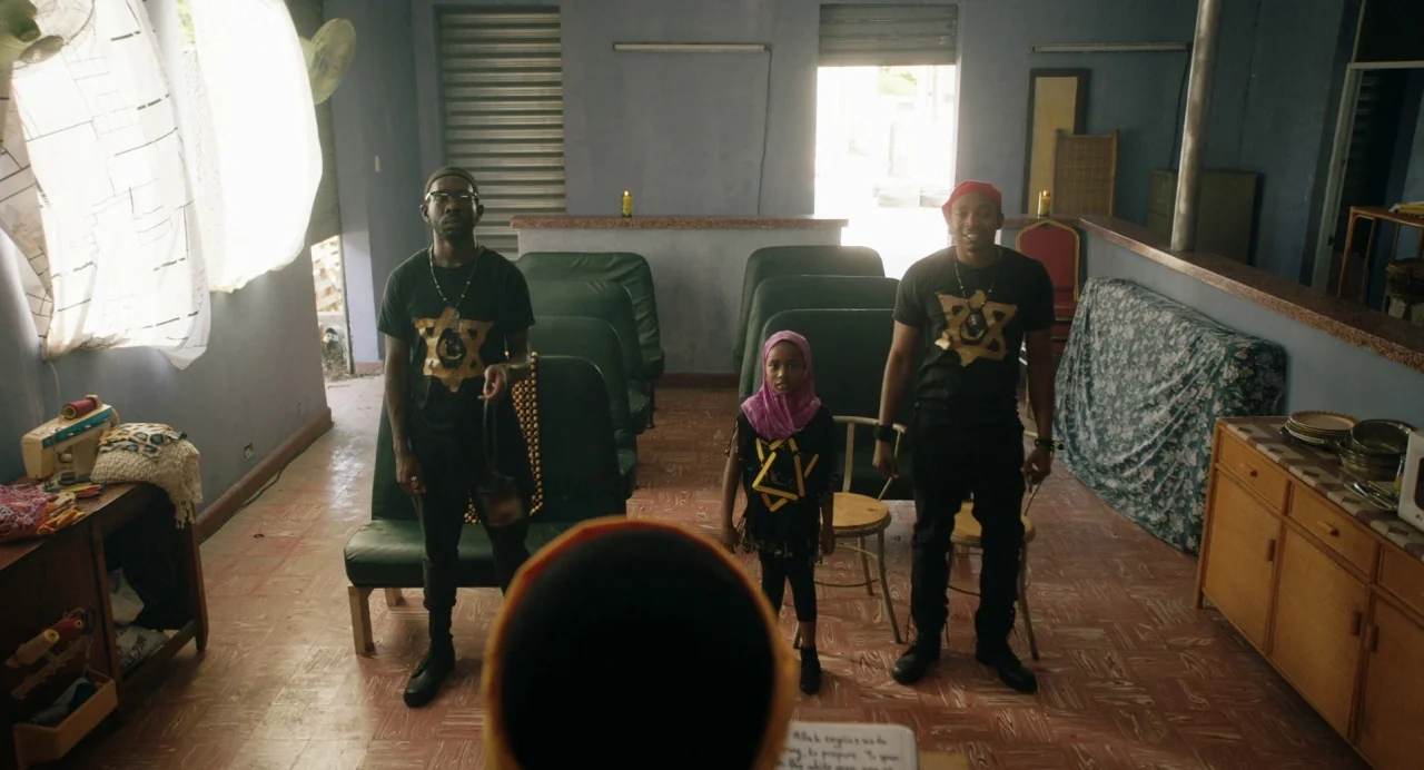 The Day Shall Come (2019) movie still: wide — A spare community room features rows of green plastic chairs and tiled floors. Three individuals stand…