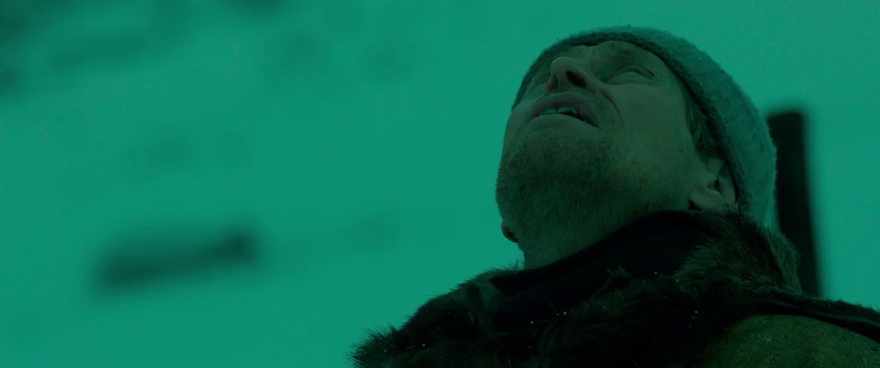 Siberia (2019) movie still: closeup — A man stands amidst a featureless, snowy expanse beneath a dim, uniformly green-tinted sky. He looks directly…