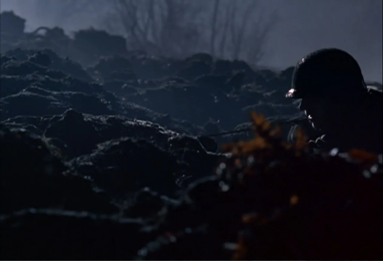 When Trumpets Fade (1998) movie still: medium — A nighttime scene set in a muddy trench with dark, piled earth banks. A soldier in a helmet hunkers…