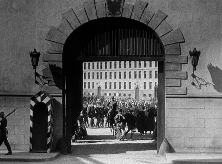 All Quiet on the Western Front (1930) movie still: wide — A stone archway frame leads to a courtyard filled with soldiers marching in formation. A…