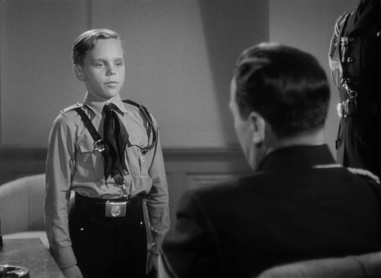 To Be or Not To Be (1942) movie still: medium — An interior office with plain walls provides the setting for a formal interaction. A young boy in…