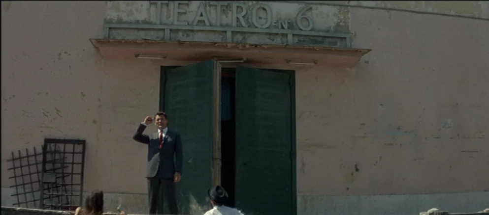 Contempt (Le Mépris) (1963) movie still: fullbody — The exterior of a weathered, pink building features a sign reading TEATRO N. 6. A man in a dark…