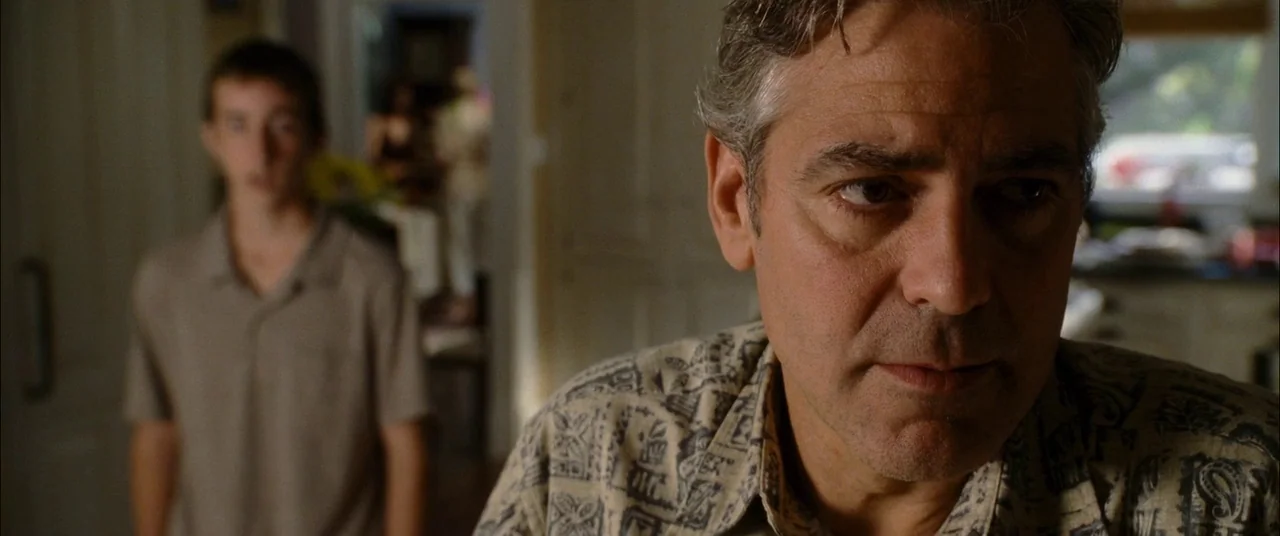The Descendants (2011) movie still: closeup — A man stands in the foreground of a softly lit residential interior. A teenager stands out of focus in…
