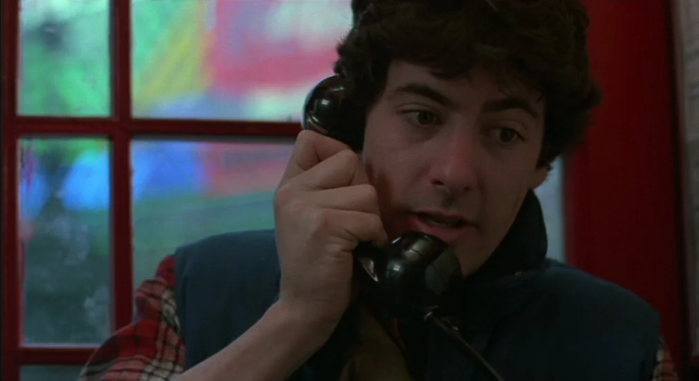 An American Werewolf in London (1981) movie still: closeup — A man stands inside a classic red glass-paneled telephone booth during the day. He…