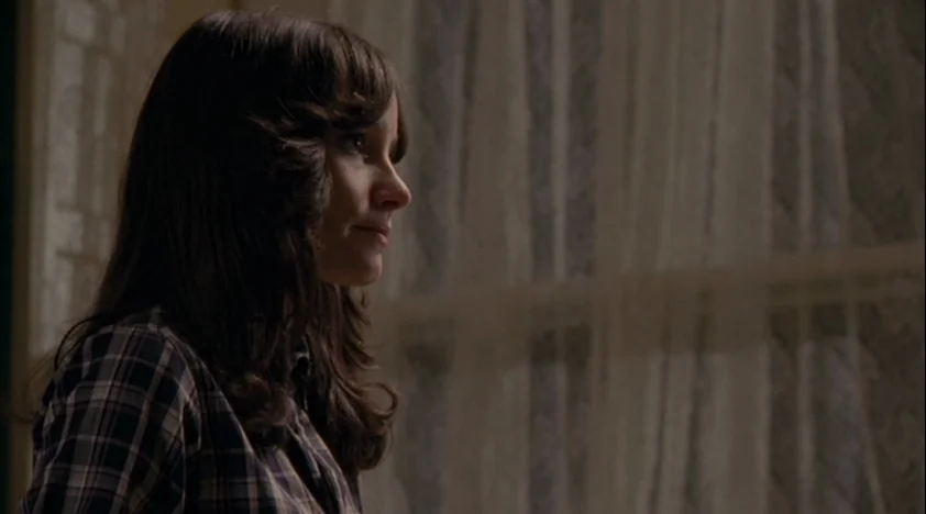 The House Of The Devil (2009) movie still: medium — An interior space with a window covered by sheer curtains in the background. A woman with long…