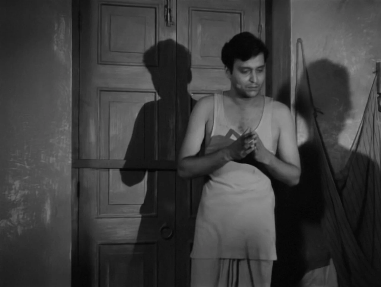 The Coward (1965) movie still: medium — An interior space with a wooden door and textured walls casting long harsh shadows. A man in a simple vest…