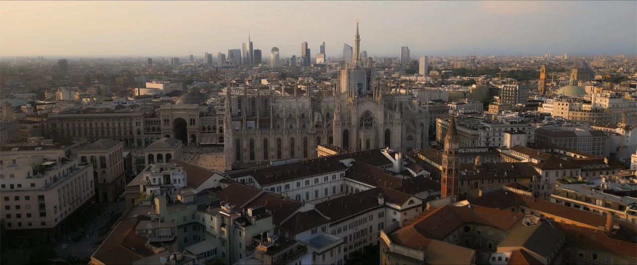 Love Me Love Me (2026) movie still: establishing — A wide aerial view shows the historic Milan Cathedral surrounded by dense urban buildings. Modern…