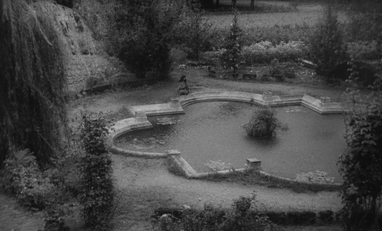 The Wild Child (1970) movie still: wide — A quiet park with a clover-shaped stone pond and overgrown foliage. A single woman sits on the rim of the…