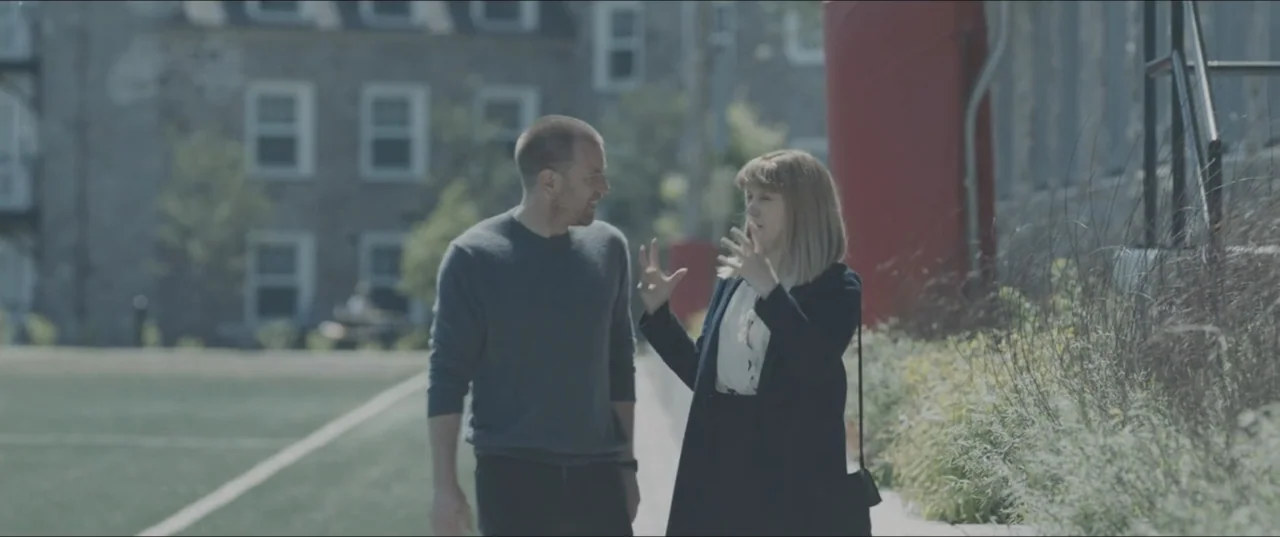 Zoe (2018) movie still: medium — A man and woman walk together along an outdoor path beside an artificial field. They are talking to each other…