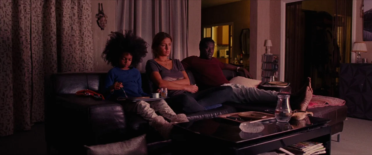 The Five Devils (2022) movie still: wide — A dark, furnished space features a large dark couch occupied by three people. They sit together facing…