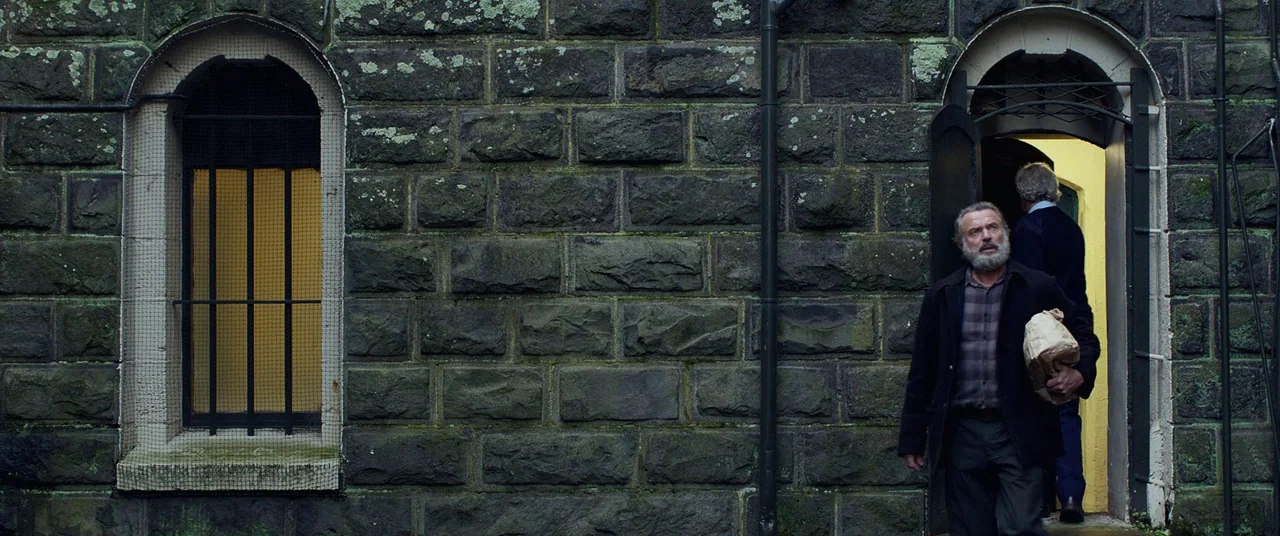 Hunt for the Wilderpeople (2016) movie still: medium — A dark, textured stone exterior wall features a barred arched window and a doorway. A bearded…