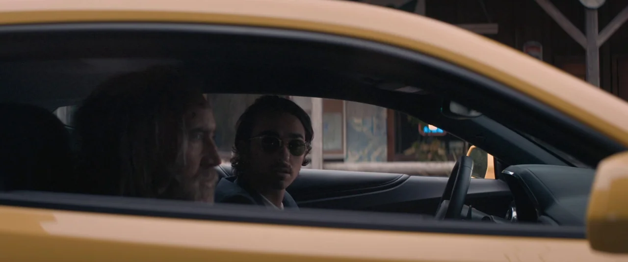 Pig (2021) movie still: medium — A yellow car sits on a street with a blurred storefront visible in the background. Two men are seated inside the…