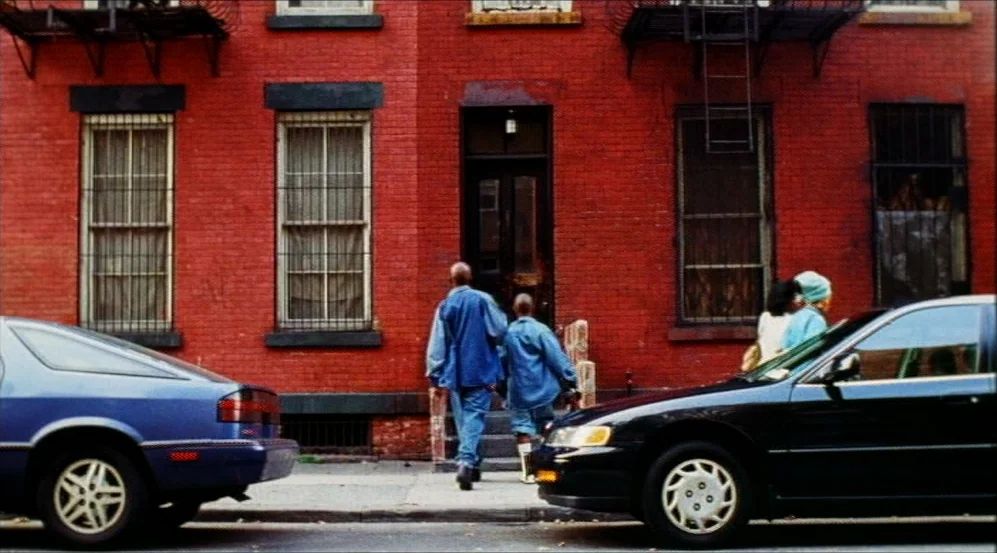 Clockers (1995) movie still: wide — A red brick residential building features black window frames and a central doorway. Two men walk toward the…