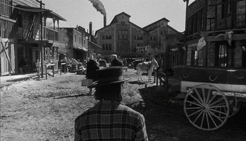 Dead Man (1995) movie still: medium — A dusty dirt road runs through a 19th-century western town with wooden buildings. A man in a top hat stands in…