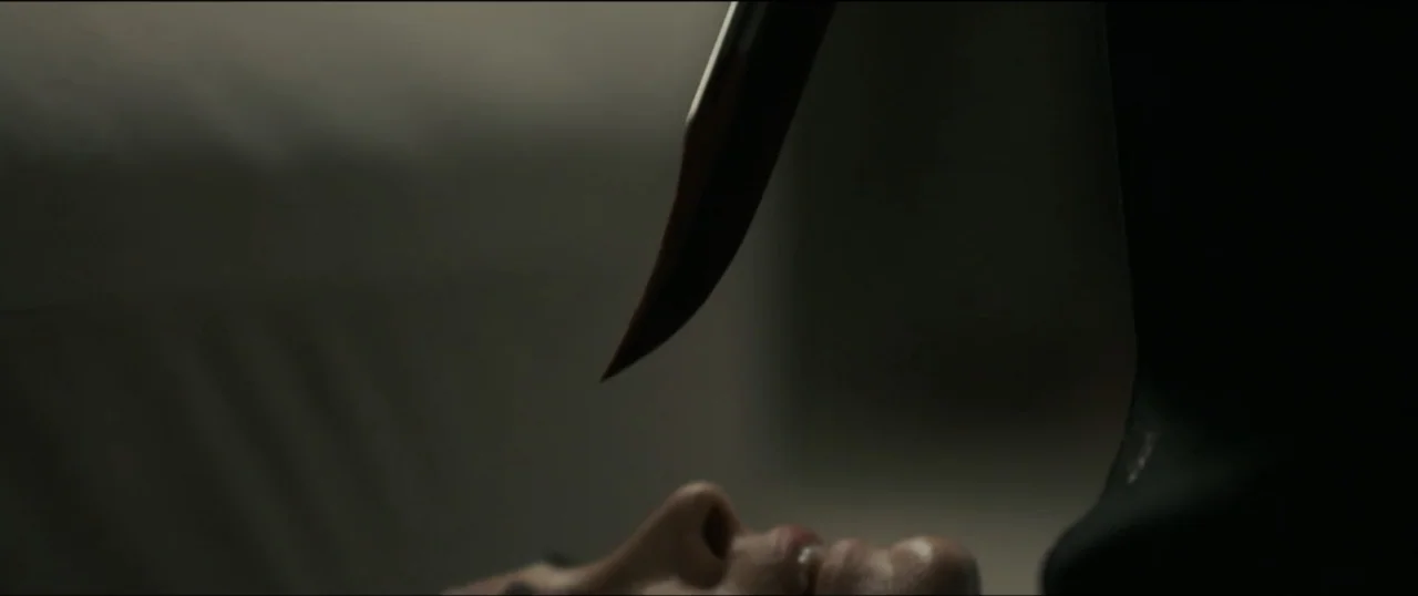 Scream VI (2023) movie still: closeup — A dark, nondescript indoor space features an out-of-focus face lying on a surface. A sharp knife blade…