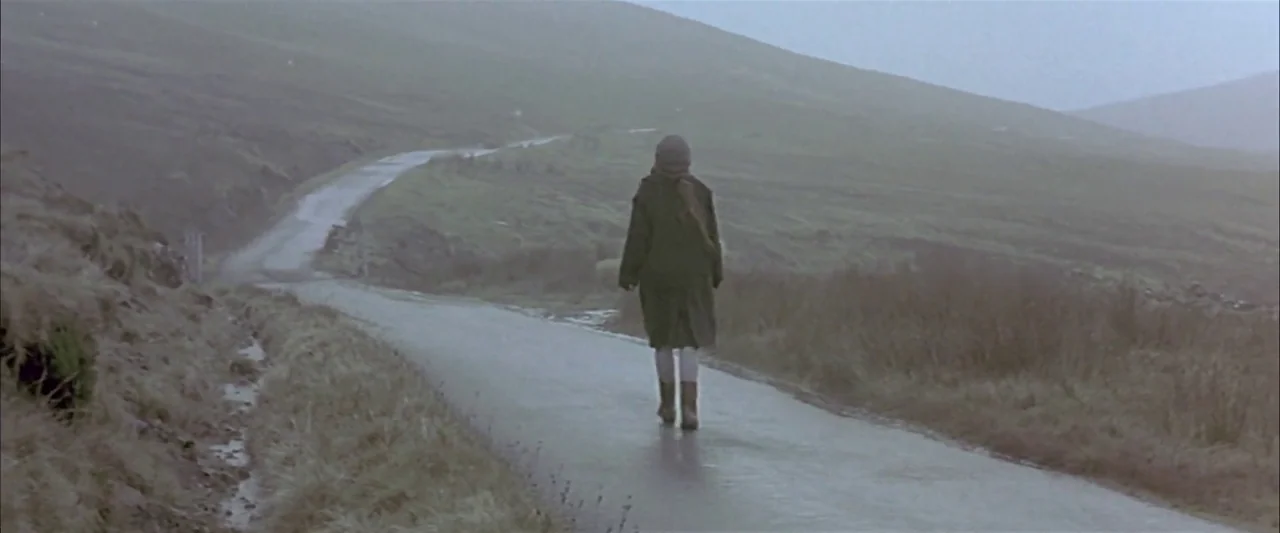 Breaking the Waves (1996) movie still: fullbody — A wet asphalt road winds through a foggy, barren hilly landscape under grey skies. A lone person…