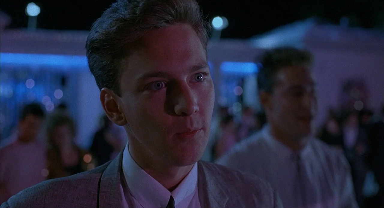 Less Than Zero (1987) movie still: closeup — A dimly lit indoor space features blurred dance lights in the background. A young man in a gray suit…