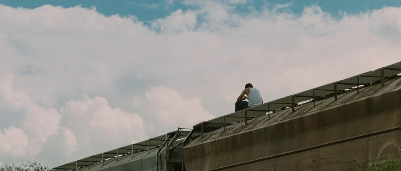 Sin Nombre (2009) movie still: wide — A view from below shows a large rusted metal freight train car roof. A lone male passenger sits on the top…