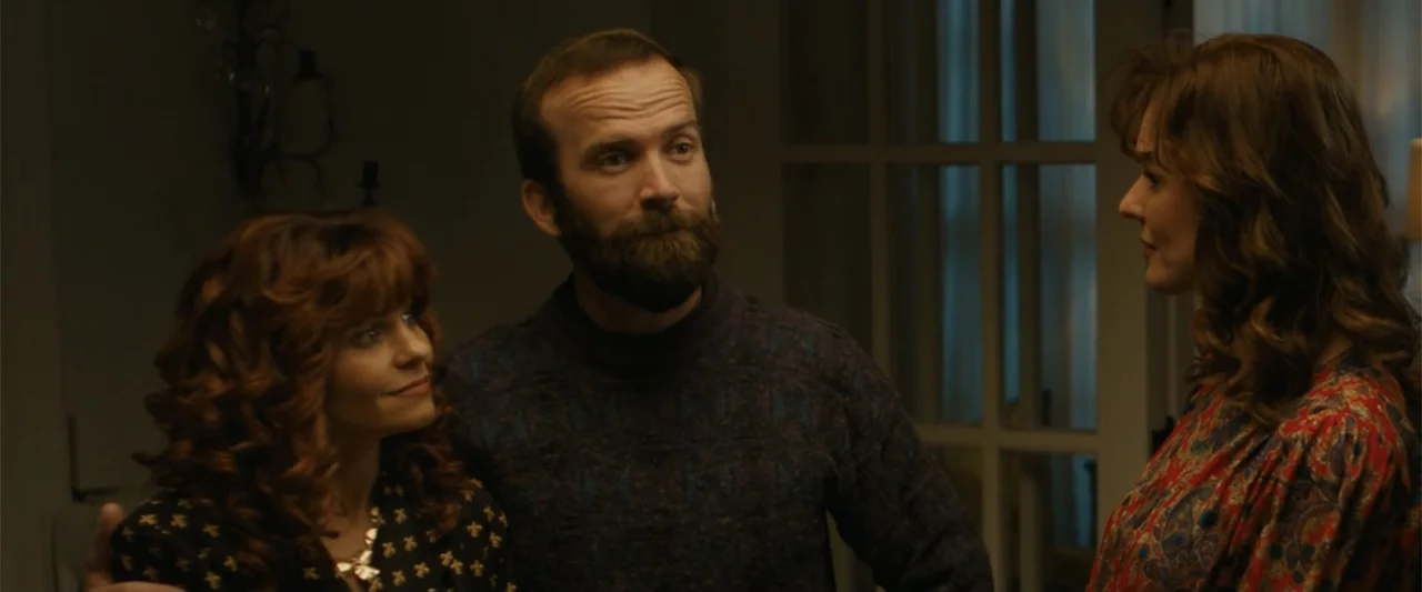 Unsung Hero (2024) movie still: medium — A dimly lit indoor space featuring a group of three adults in casual attire. Two women and one bearded man…
