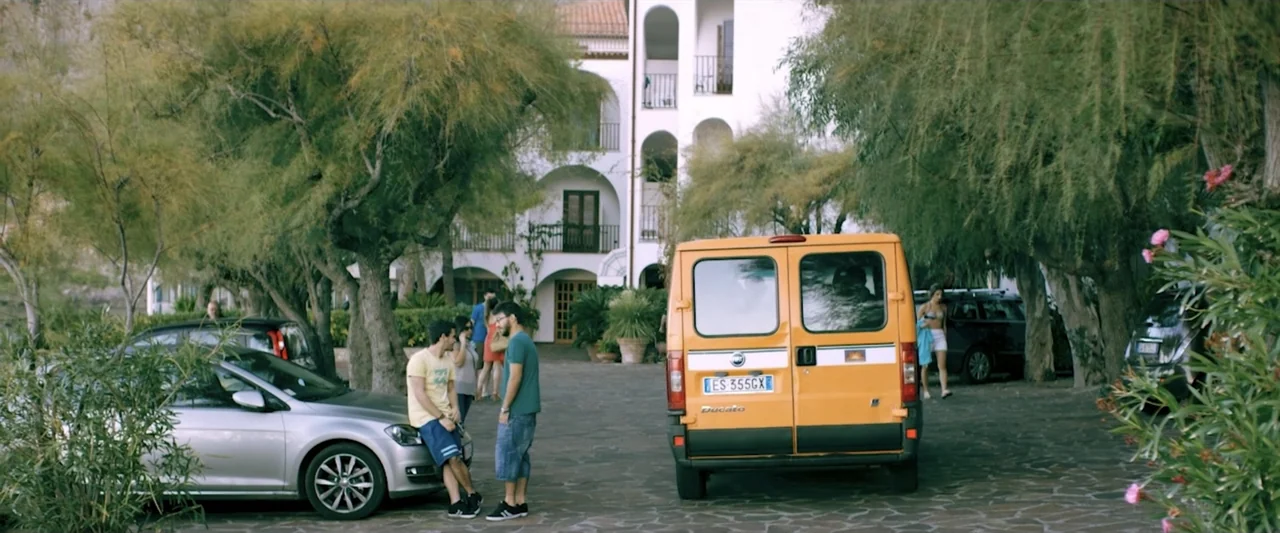 Taranta On the Road (2017) movie still: wide — A sunny Mediterranean courtyard features a white multi-story building and several parked vehicles.…