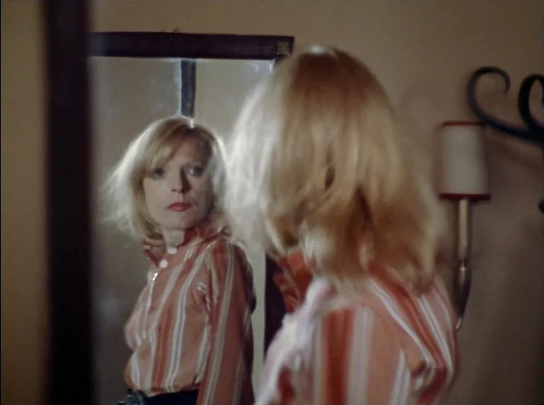 Fear Of Fear (1975) movie still: medium — A woman looks into a wall-mounted mirror inside a minimally furnished room. The woman views her reflection…