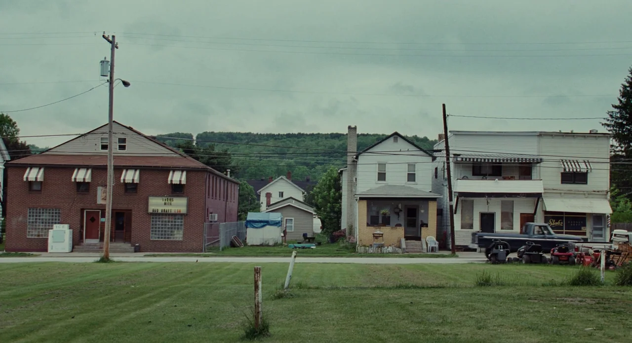 Promised Land (2012) movie still: wide — A quiet small-town street features a brick building, a white house, and shops. Several lawn mowers are…