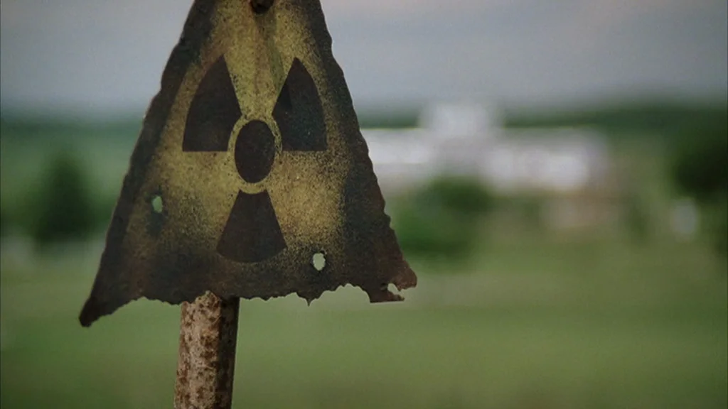 Everything Is Illuminated (2005) movie still: closeup — A weathered, triangular radiation warning sign sits in the foreground of an open field. In…