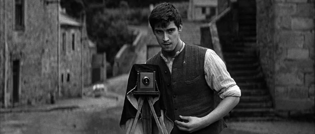 Jude (1996) movie still: medium — An old stone village street features traditional masonry houses and a stone staircase. A young man stands behind a…