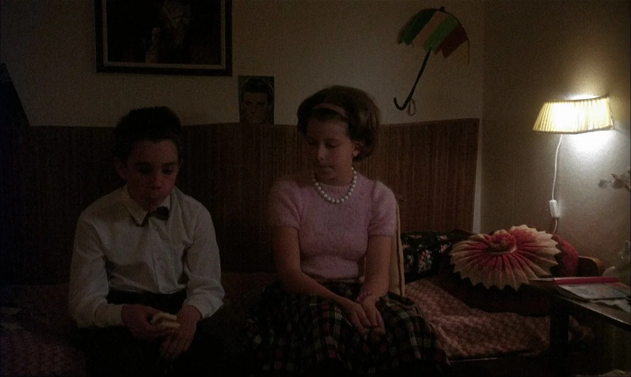 My Life As A Dog (1985) movie still: medium — A bedroom wall features a framed picture and a decorative umbrella wall hanging. A boy and a girl sit…