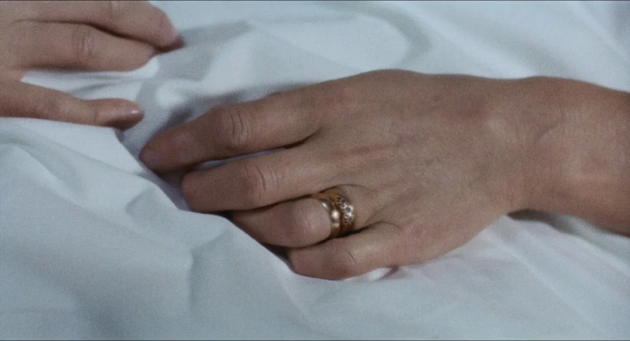 The Touch (1971) movie still: closeup — A pair of hands with rings rests on crumpled, smooth white bed sheets. The surface is textured by folds in…