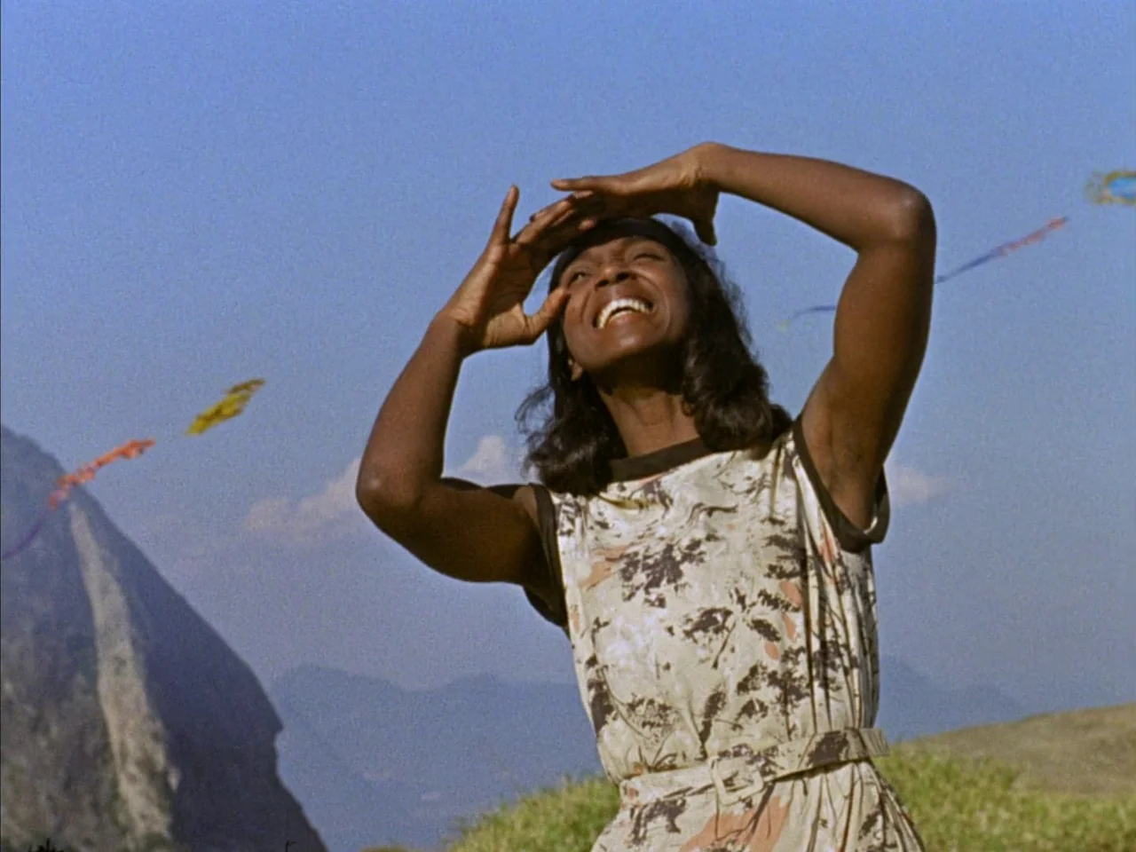 Black Orpheus (1959) movie still: medium — A woman stands on a grassy slope with large rocky mountains in the background. She looks toward the sky…