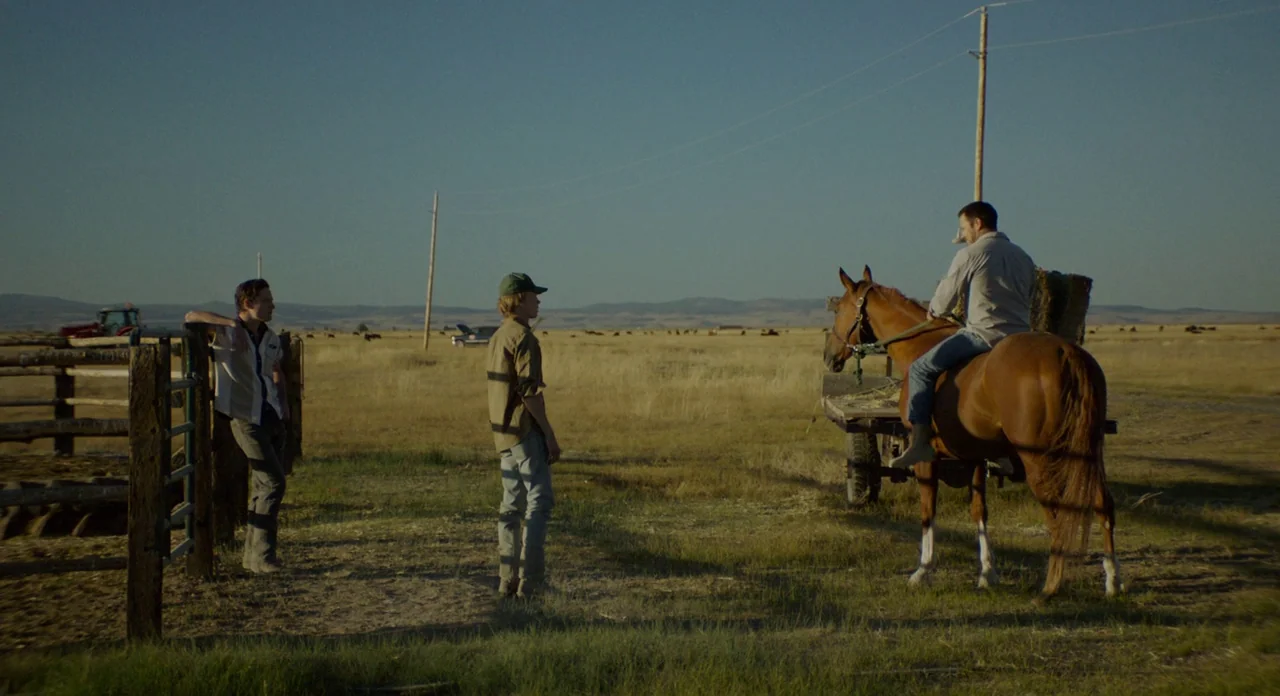 Lean on Pete (2017) movie still: wide — An open rural landscape features a wooden fence and a dry grassy field. Three men stand together in the…