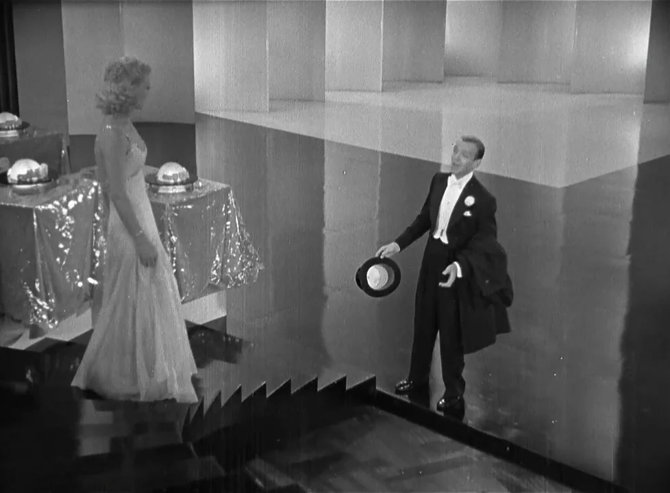 Swing Time (1936) movie still: fullbody — A spacious ballroom with a highly polished floor features large geometric white pillars. The man stands on…