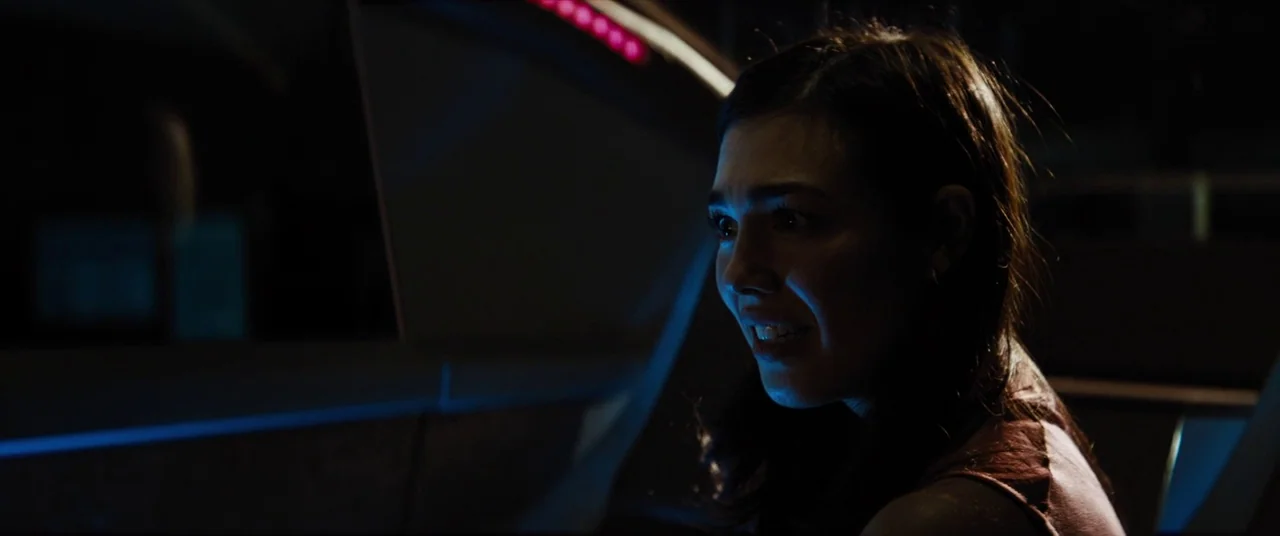 Upgrade (2018) movie still: closeup — A dark, cramped interior of a vehicle at night with blue light reflection visible. A young woman with her…