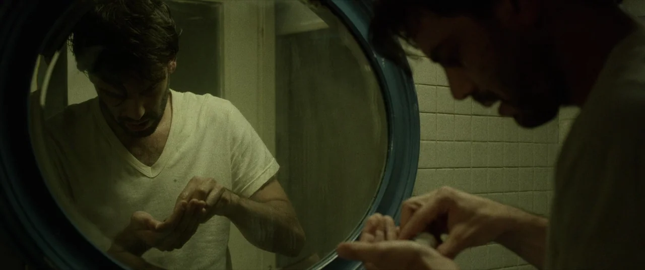 Terrified (2017) movie still: medium — A dimly lit tiled bathroom features a large circular mirror mounted on the wall. A man in a plain white…