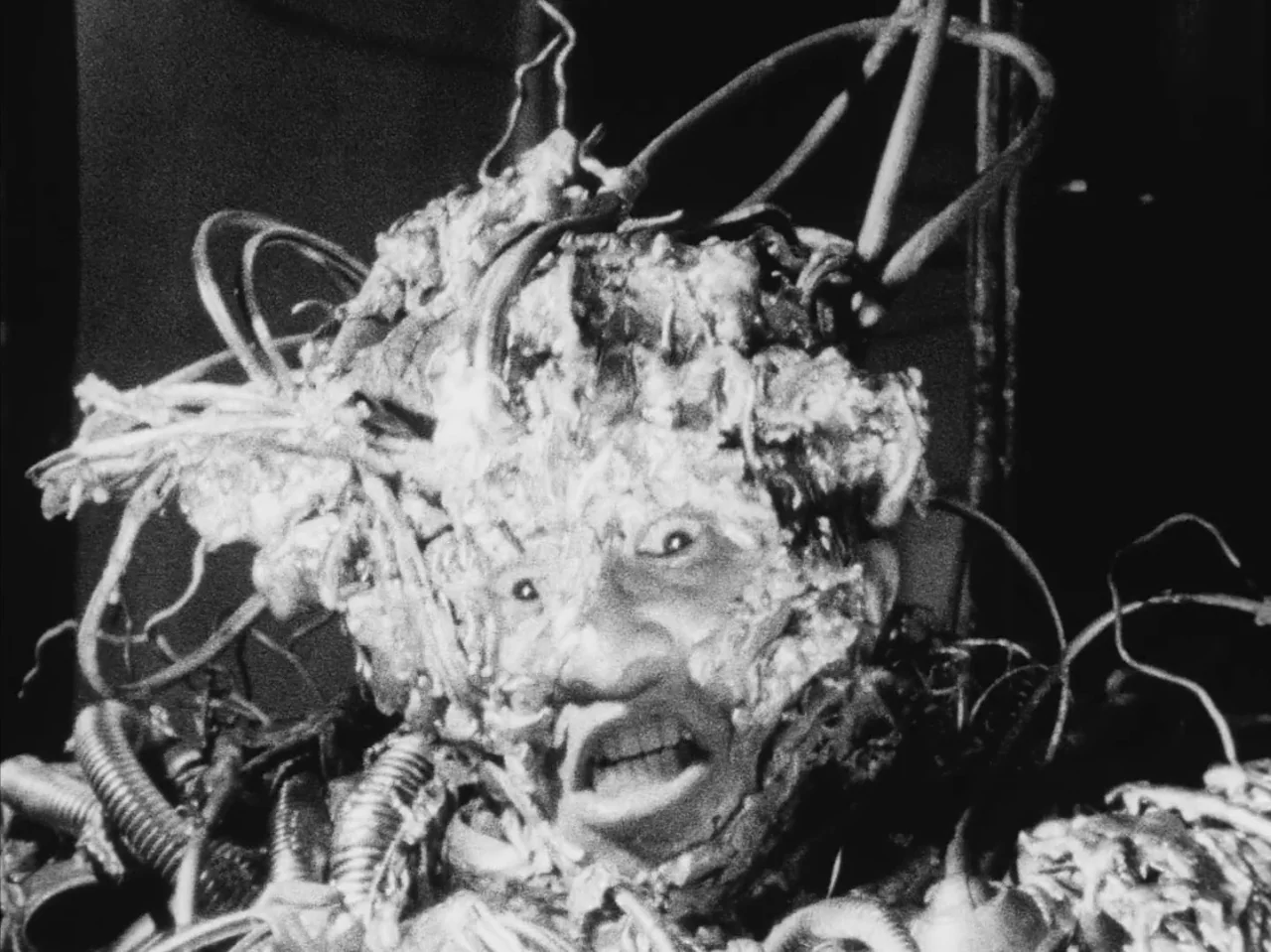 Tetsuo: The Iron Man (1989) movie still: closeup — A man is entwined with wires and organic matter inside a dark, confined space. His face is…