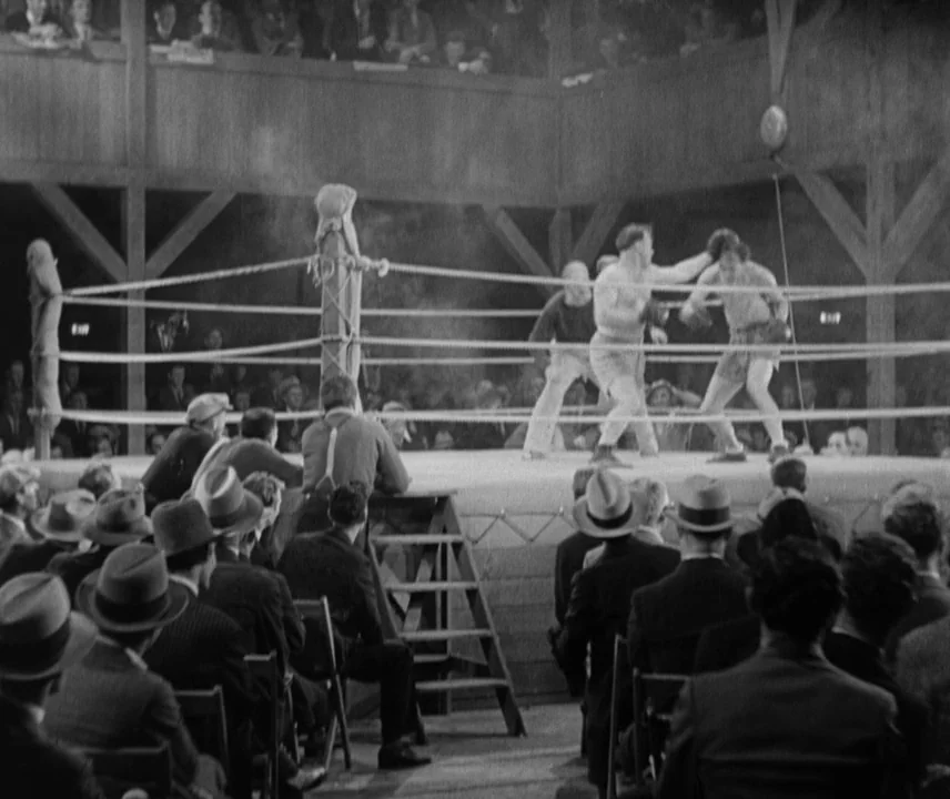 City Lights (1931) movie still: wide — A rustic boxing ring surrounded by spectators seated on wooden chairs. Two fighters engage in a bout while a…