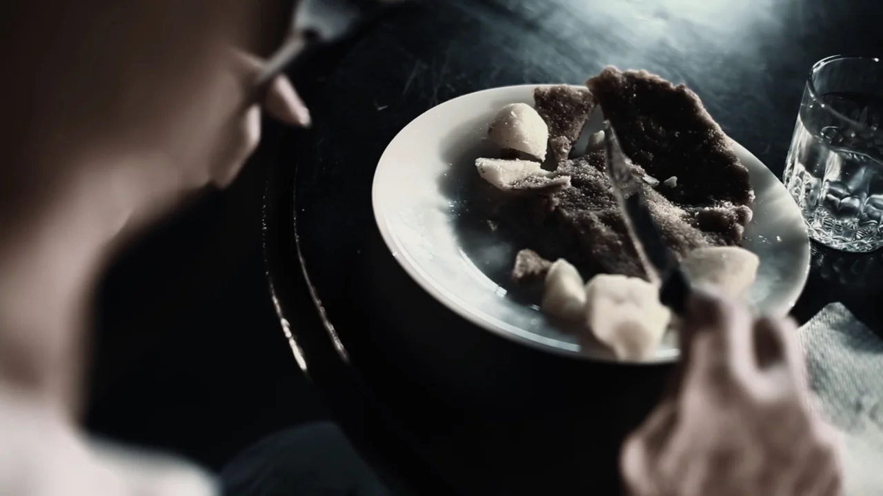 Still Life (2013) movie still: closeup — A dark dining table holds a white plate containing sliced food and a glass. A person is seen from behind…