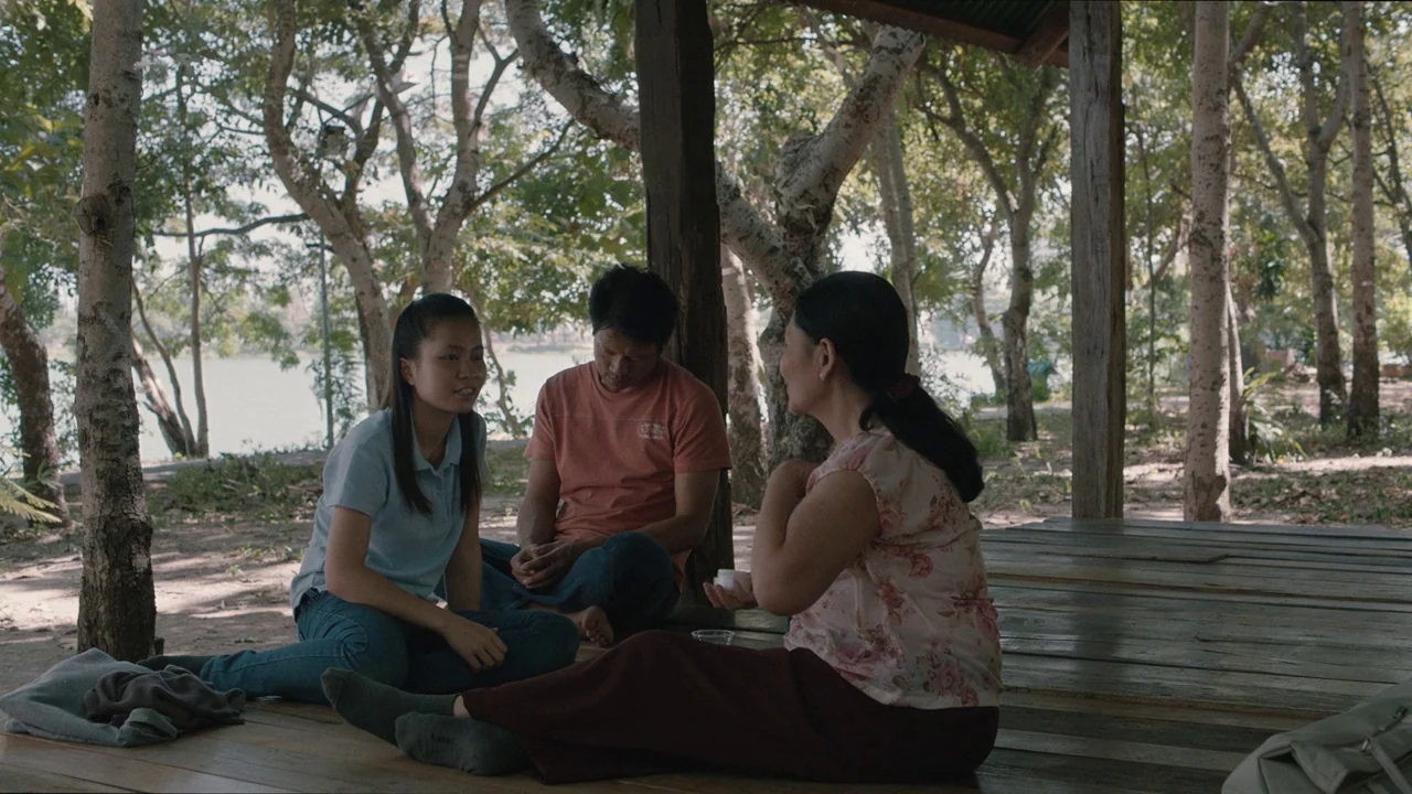 Cemetery of Splendour (2015) movie still: fullbody — An open-air wooden platform sits amidst a grove of trees near a riverbank. Three people sit on…