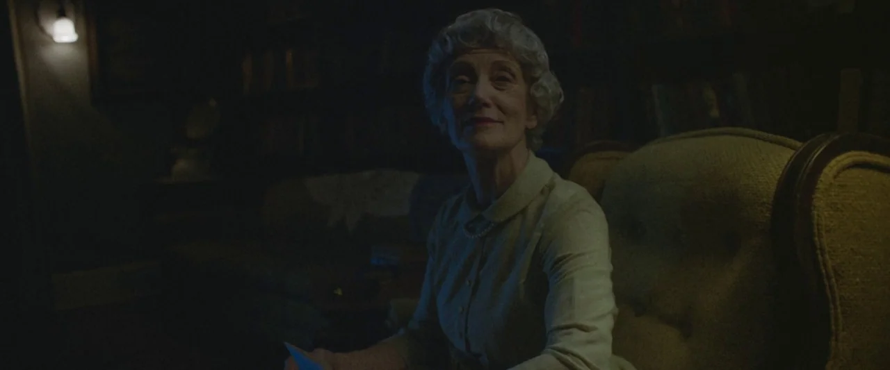 The Vast of Night (2019) movie still: medium — A dim room featuring bookshelves and a large fabric-upholstered armchair in the shadows. An elderly…