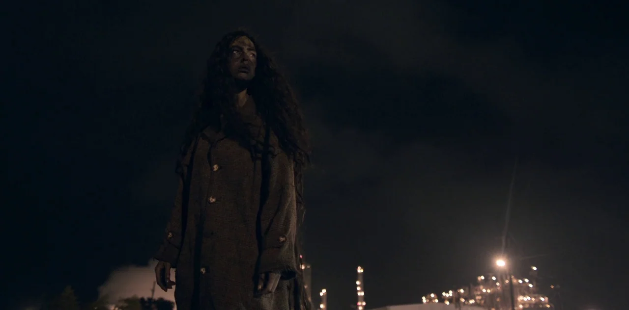 Darlin’ (2019) movie still: medium — An industrial facility at night features illuminated refinery stacks in the blurred background. A woman with…