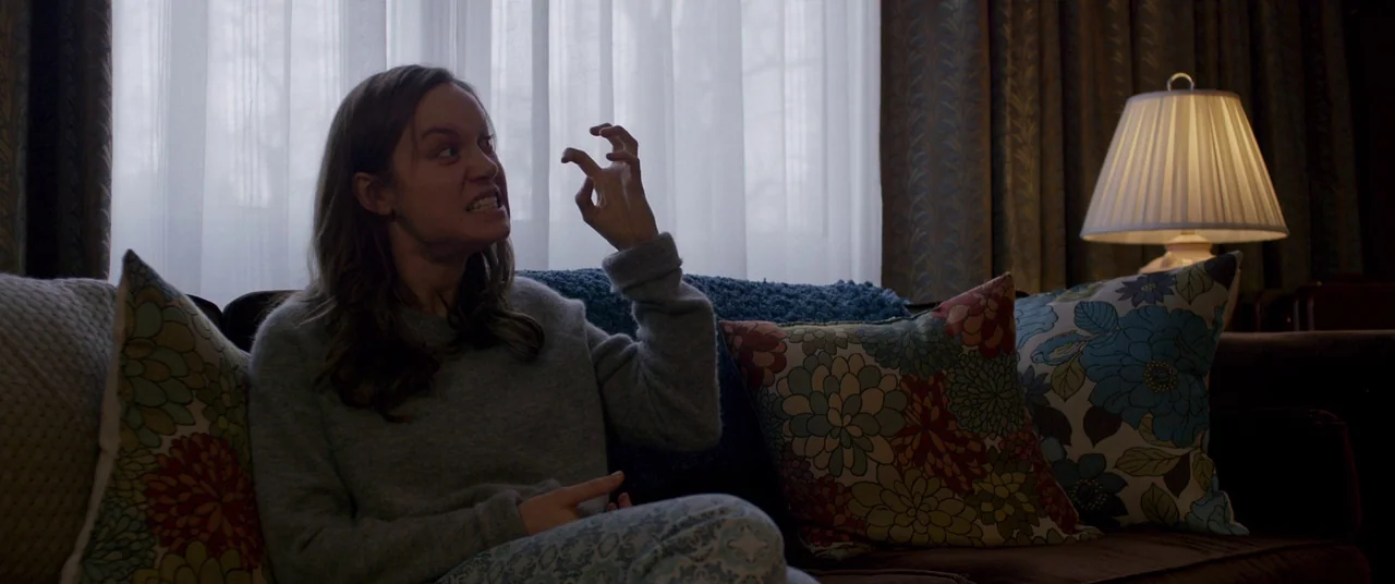 Room (2015) movie still: medium — A domestic living room with a brown couch and floral patterned throw pillows. A woman sits on the couch and holds…