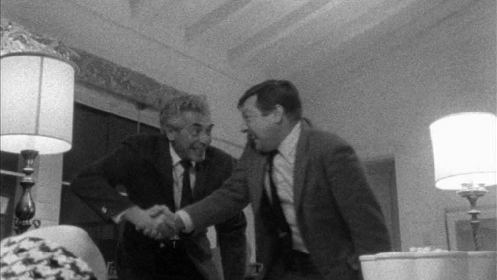Faces (1968) movie still: medium — A well-lit room interior features two men meeting in a formal setting. The men exchange a handshake while facing…