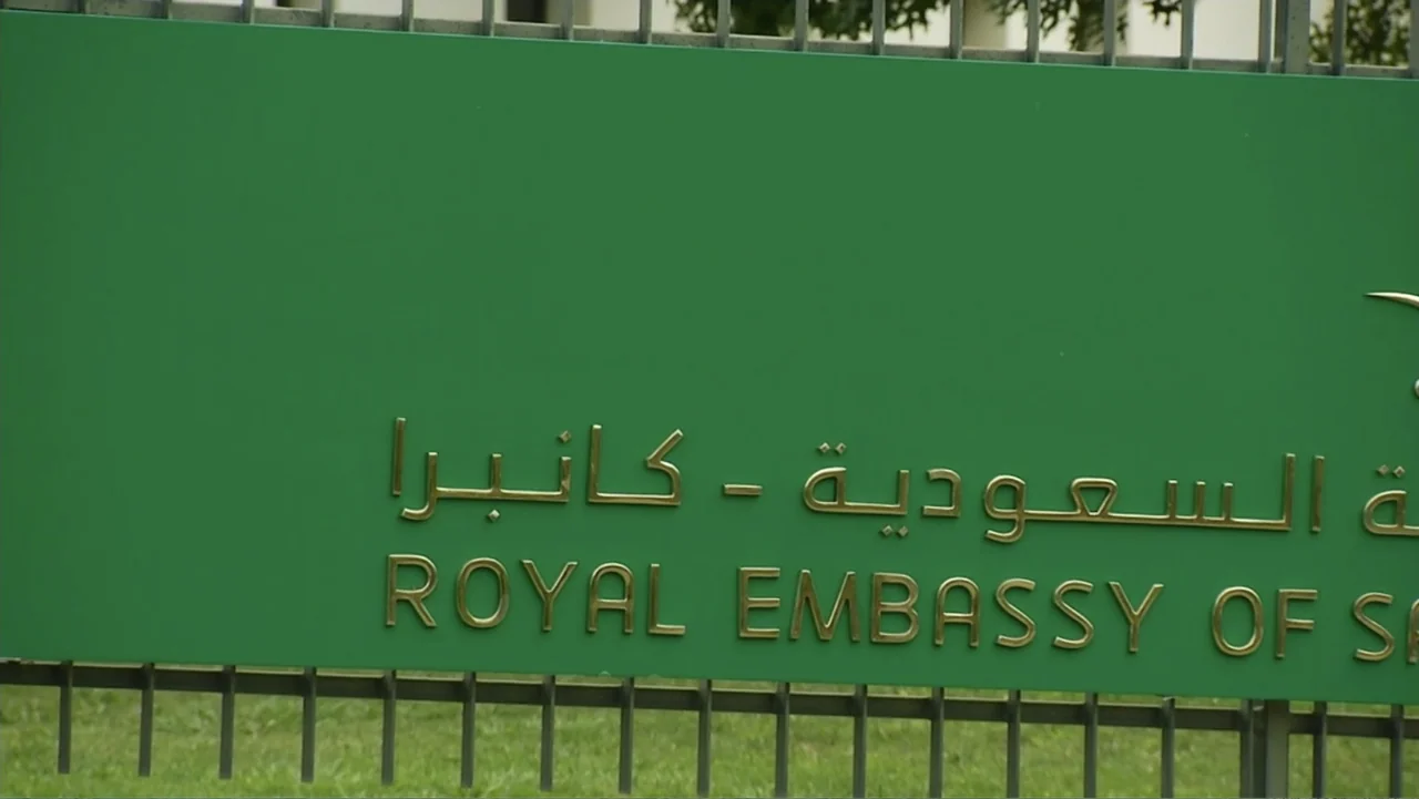 Escape From Saudi (2019) movie still: closeup — A green rectangular plaque with embossed gold lettering mounted on a metal fence. The sign displays…