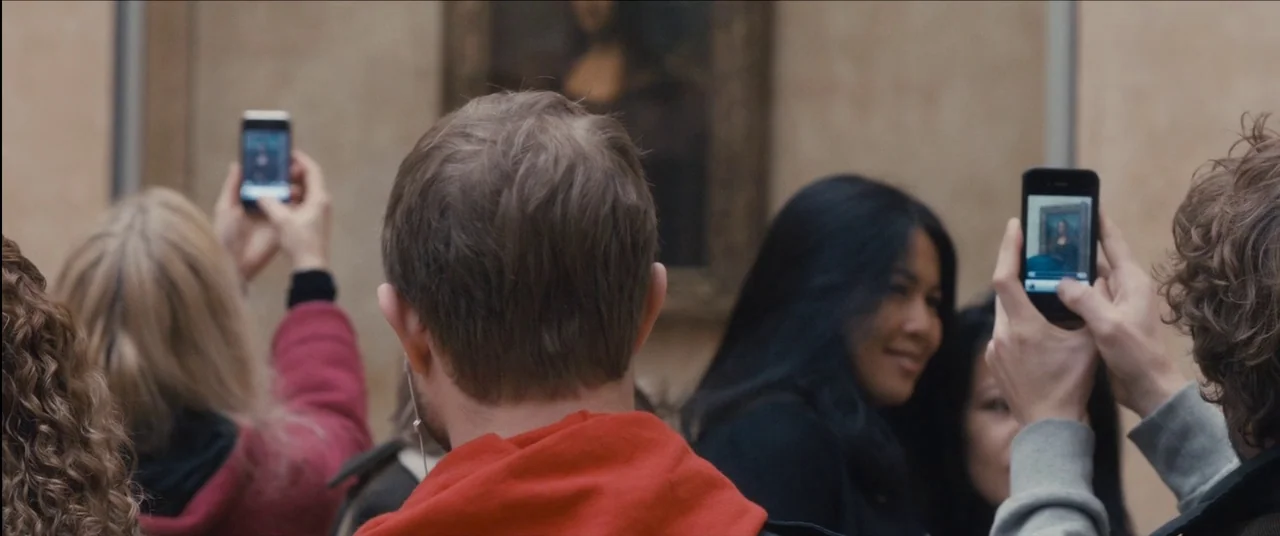 Simon Killer movie still: medium — A crowded public space with beige walls decorated with a framed painting. Several people are facing forward,…