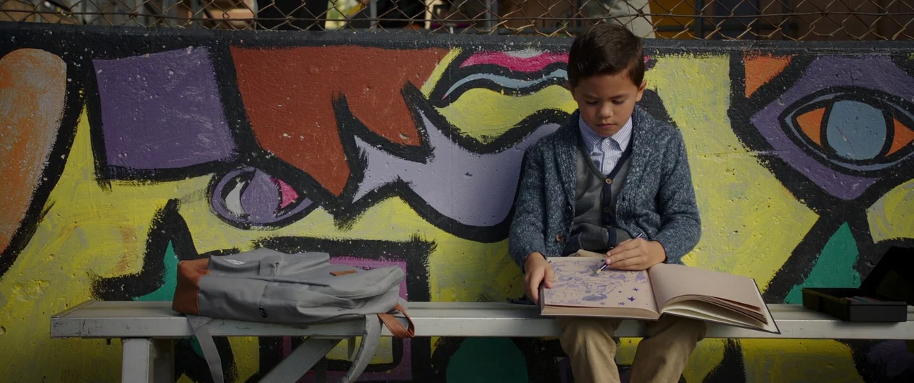 A Wrinkle In Time (2018) movie still: medium — A young boy sits on a metal bench in front of a colorful mural. He draws in a large sketchbook with a…