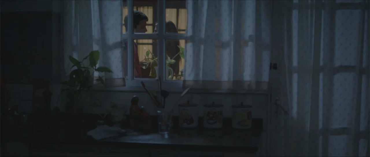Nahir (2024) movie still: medium — A dimly lit kitchen features a countertop cluttered with various containers and a plant. Two teenagers stand…